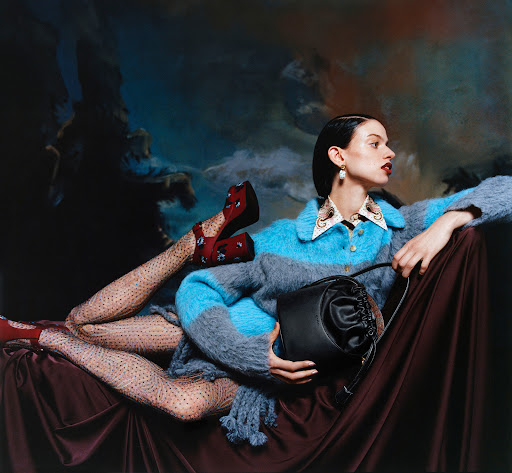 A woman lies on a draped surface wearing a blue and grey sweater, patterned tights, red platform shoes, and carrying a black bag.