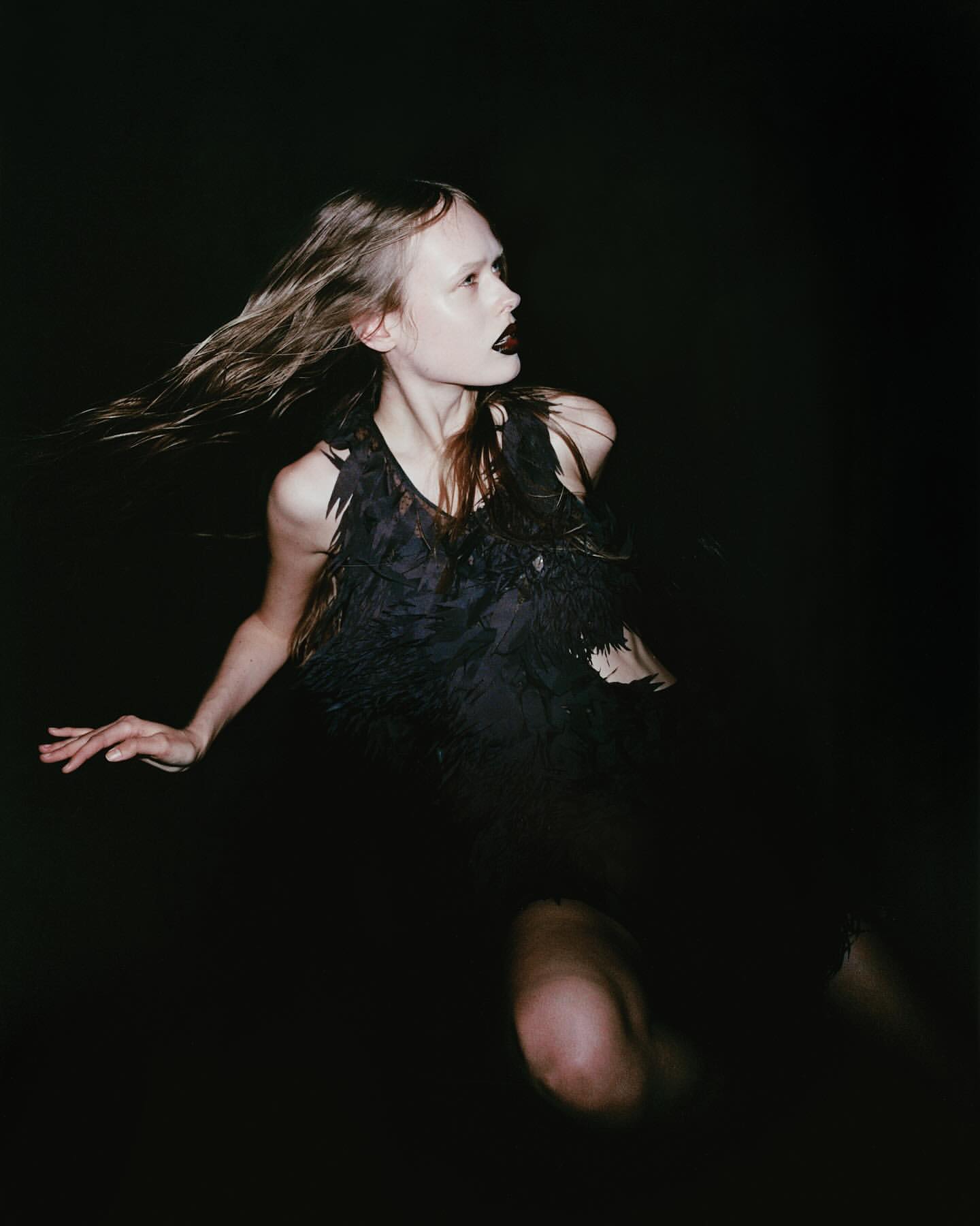 A woman with windblown hair and dark lipstick wears a black feathered top against a dark background in a dramatic portrait.
