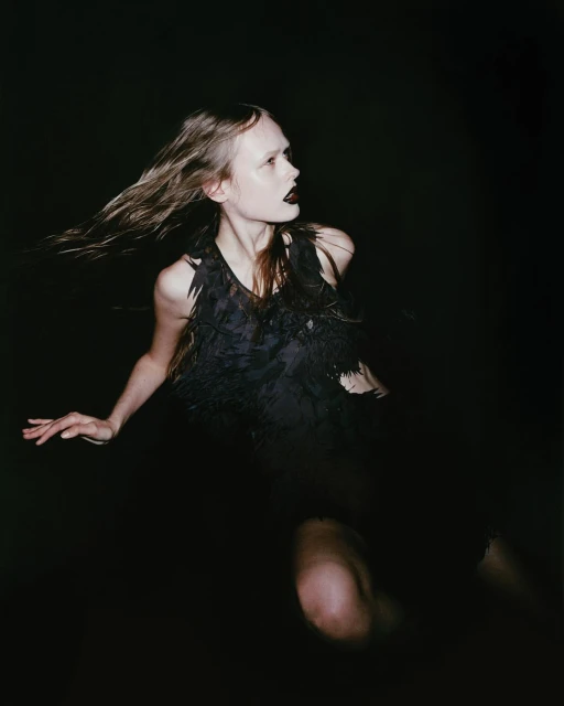 A woman with windblown hair and dark lipstick wears a black feathered top against a dark background in a dramatic portrait.
