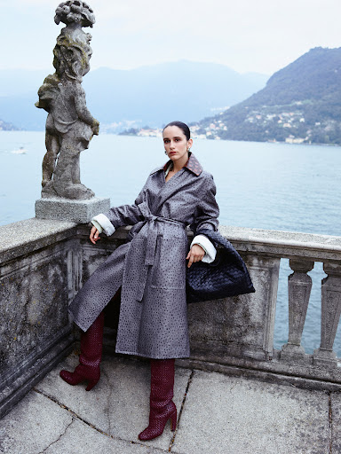 Woman in patterned coat and boots poses on a stone balcony overlooking Lake Como, Italy, with mountains and greenery in the background.