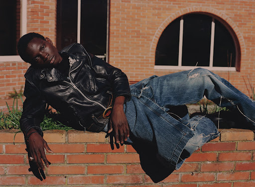 A Black male model wearing a black leather jacket and ripped jeans poses on a brick ledge with a brick building background.