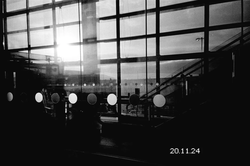 Black and white photo of a large window with a grid pattern, reflecting an outdoor scene with stairs and a distant light.