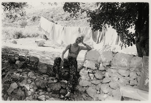 Black-and-white shot of a young woman in printed top and pants sitting on a stone wall in a rural setting with clotheslines in the background.