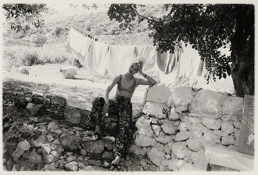 Black-and-white shot of a young woman in printed top and pants sitting on a stone wall in a rural setting with clotheslines in the background.