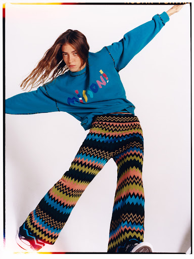 Fashion shot of a woman in a turquoise top and multicolored pants in a dynamic pose against a white backdrop in a studio setting.