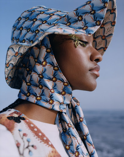 Profile of an African-American woman wearing a floral hat and scarf, styled with natural greenery across her eye, with ocean backdrop.
