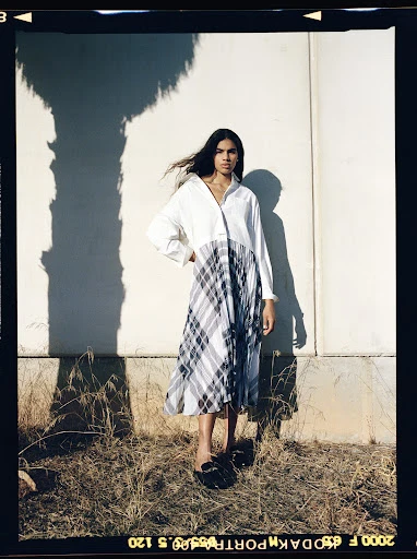 A woman stands outdoors in a white top and grey plaid dress, with long dark hair, posing against a white wall with a tree shadow.