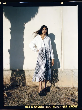 A woman stands outdoors in a white top and grey plaid dress, with long dark hair, posing against a white wall with a tree shadow.