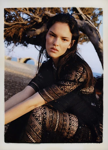 Portrait of a woman with dark hair wearing a black lace outfit, posing outdoors near a tree and the ocean on a sunny day.