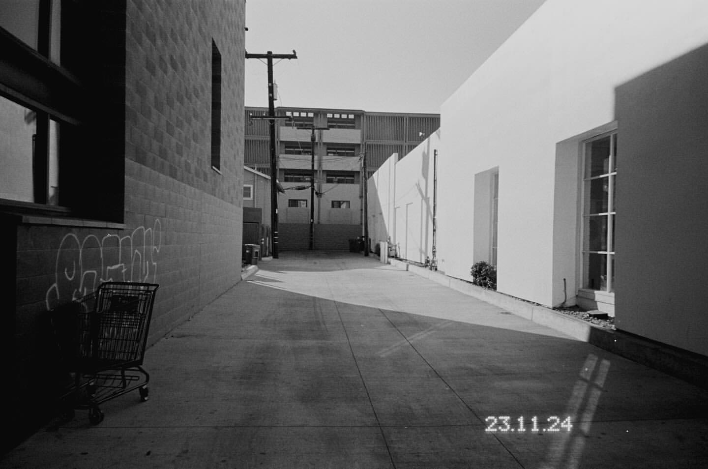 Black and white photograph of a concrete alleyway between buildings in an urban setting with a shopping cart and graffiti.