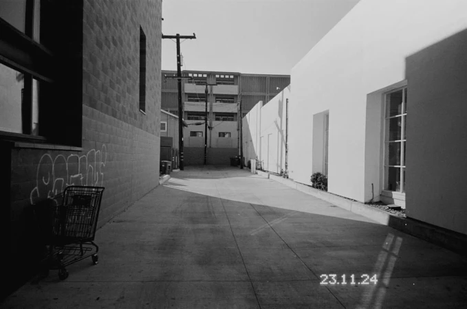 Black and white photograph of a concrete alleyway between buildings in an urban setting with a shopping cart and graffiti.