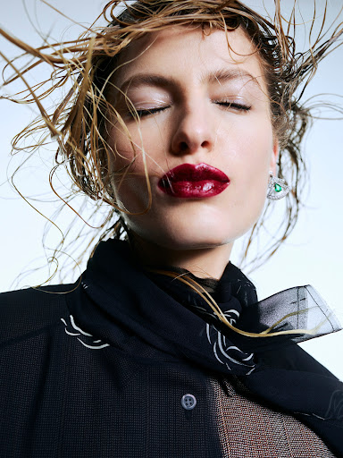 Close-up portrait of a woman with wet blonde hair blowing across her face, dark glossy lipstick, and a black button-down shirt.