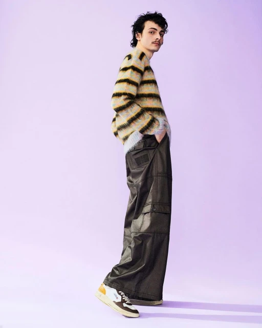 A young man with a mustache poses in a fuzzy striped sweater and wide-leg pants against a solid lavender studio background. 