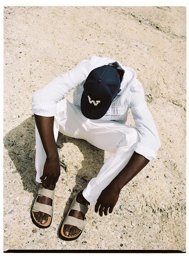 An adult man in a hooded shirt and white pants sits on the ground wearing sandals and a baseball cap. Shot from above outdoors.