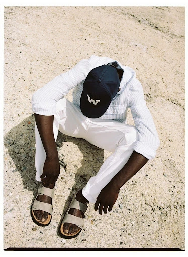 An adult man in a hooded shirt and white pants sits on the ground wearing sandals and a baseball cap. Shot from above outdoors.
