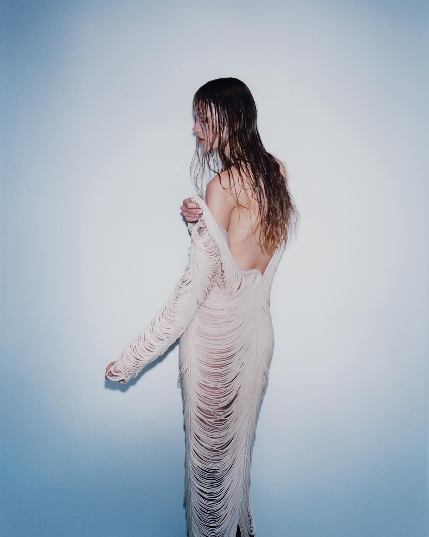 Back view of a woman in a fringed dress against a gradient blue and white background in a studio setting with soft lighting.