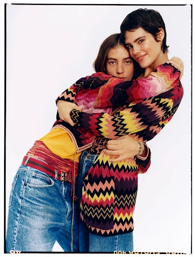 Two adult women embrace closely wearing patterned sweaters and denim in a studio setting against a plain white backdrop.