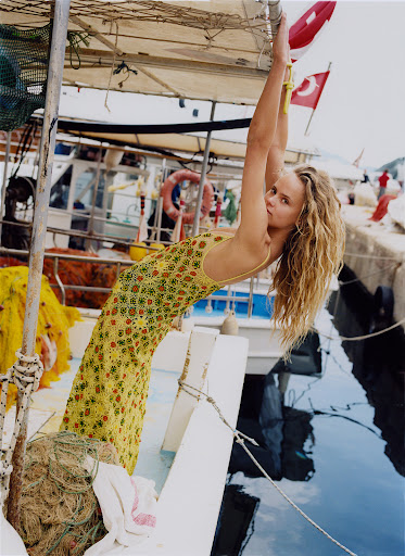 Woman in a yellow floral patterned dress hanging from the frame of a boat moored at a dock