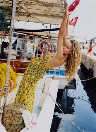Woman in a yellow floral patterned dress hanging from the frame of a boat moored at a dock