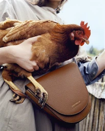 Person holding a brown chicken and wearing a tan coat with a brown leather shoulder bag by photographer Luca Campri