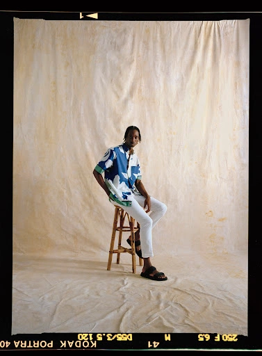 Man sitting on a bamboo stool wearing a patterned blue and white shirt and white pants against a textured backdrop