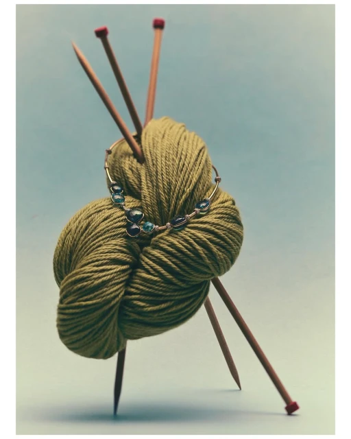 Olive green yarn ball with knitting needles and a silver and gemstone necklace on light blue background in a studio setting.