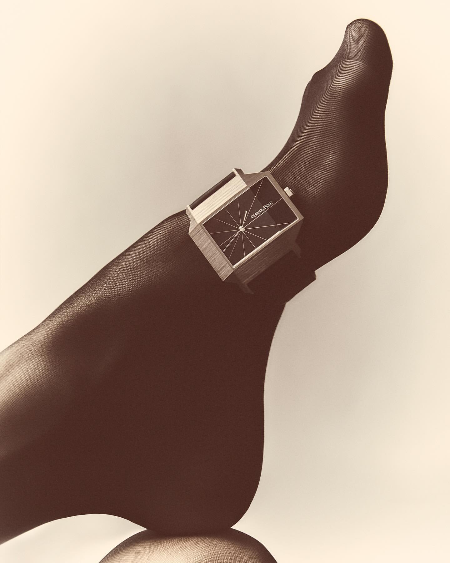 Foot with black sheer tights displaying a retro silver rectangular watch, studio shot against neutral background.