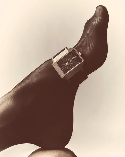 Foot with black sheer tights displaying a retro silver rectangular watch, studio shot against neutral background.