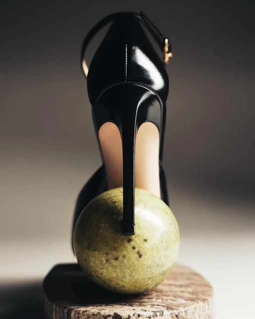 Elevated view of a glossy black high heel sandal propped on a round, light green marble, spotlighting sleek design and polished marble.