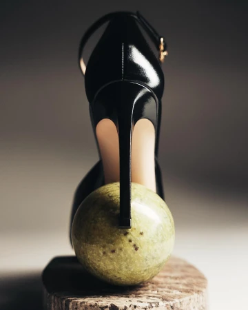Elevated view of a glossy black high heel sandal propped on a round, light green marble, spotlighting sleek design and polished marble.