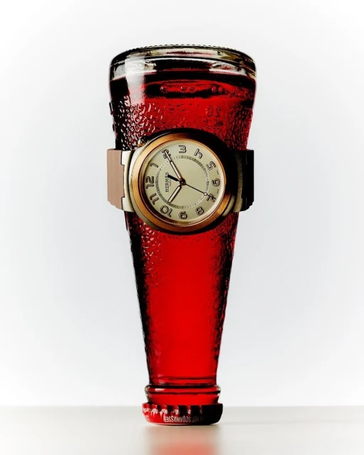 Still life studio shot of a red liquid-filled glass bottle featuring a wristwatch around its center, against a bright white background.