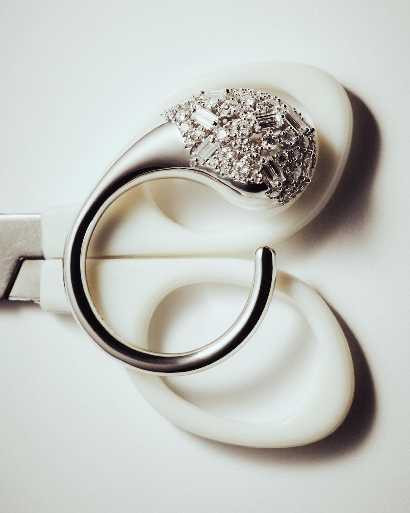 Close-up of a silver ring encrusted with diamonds, balanced inside a pair of open white craft scissors on a solid white surface.