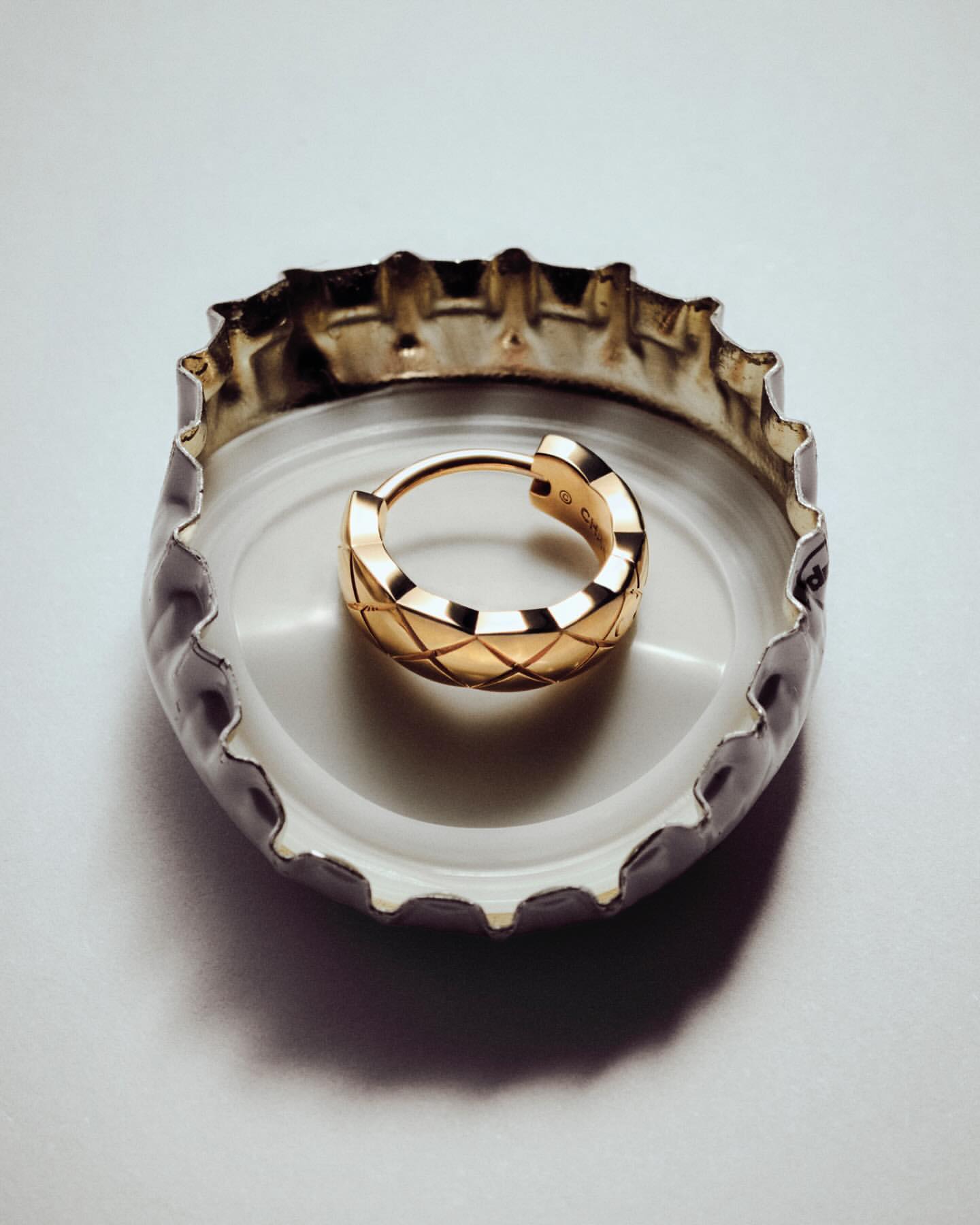 Close-up of a single small gold hoop earring with a criss-cross pattern inside a white and silver bottle cap on a white background.