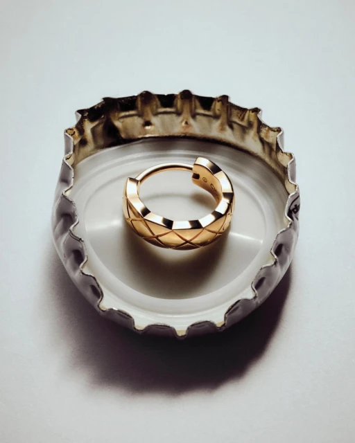 Close-up of a single small gold hoop earring with a criss-cross pattern inside a white and silver bottle cap on a white background.