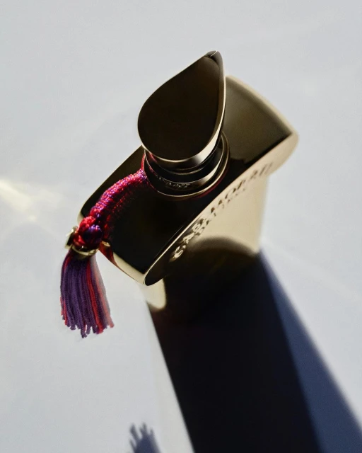 Close-up studio shot of a gold perfume bottle with a decorative tassel, casting a shadow on a white surface.