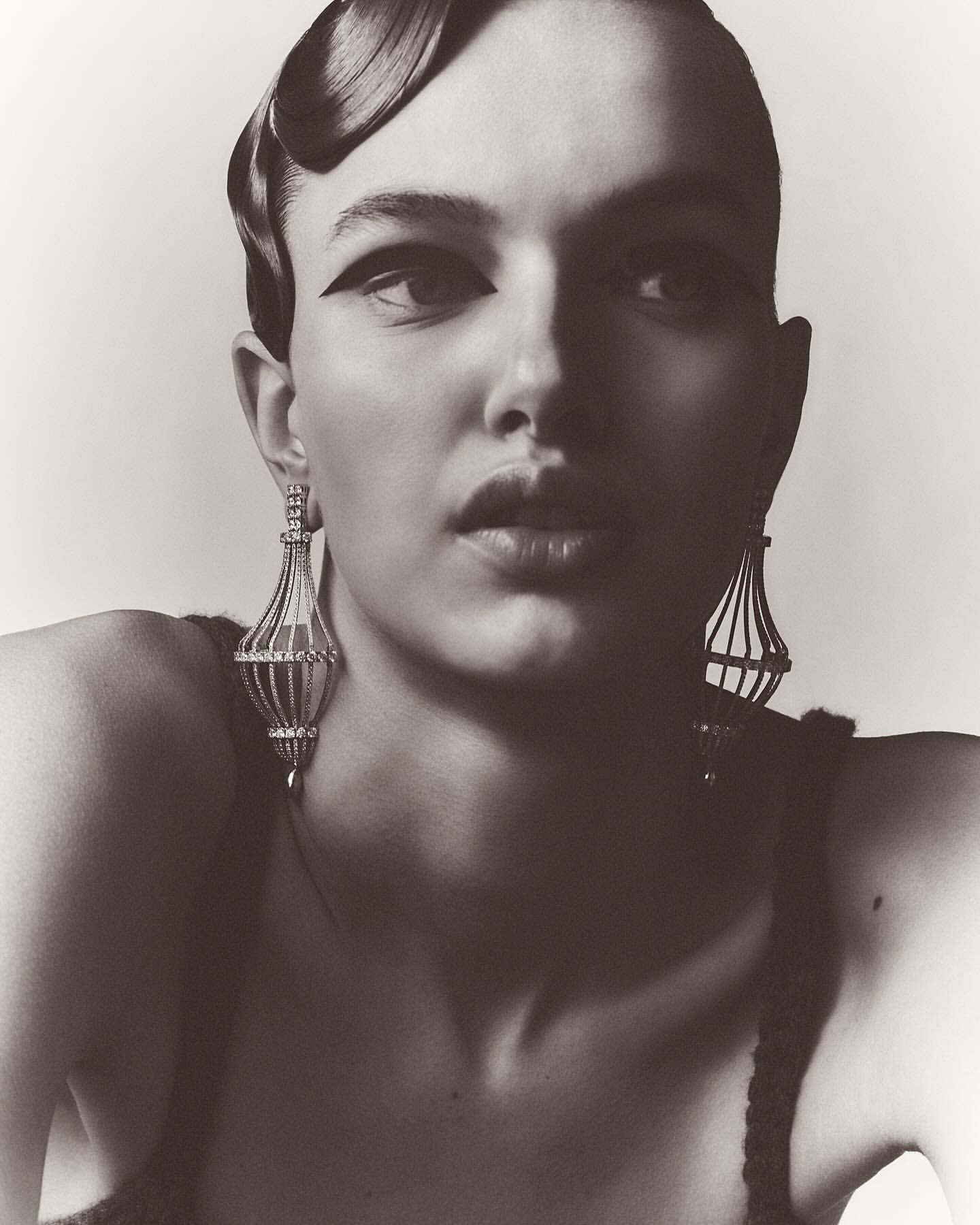 Glamorous monochrome portrait of a woman with elaborate hairstyle and jewelry, wearing black eye makeup in a neutral studio setting.