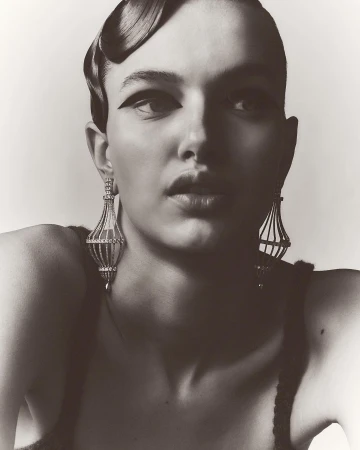 Glamorous monochrome portrait of a woman with elaborate hairstyle and jewelry, wearing black eye makeup in a neutral studio setting.