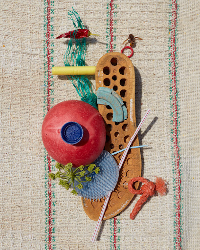Still life assemblage featuring a red ball, pepper, yellow tube, woven mat background, and a bee on top, creating a whimsical composition.