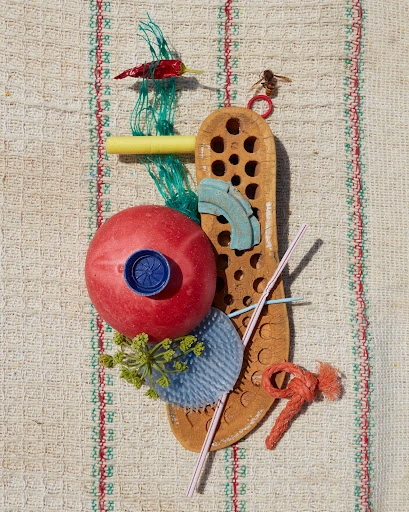Still life assemblage featuring a red ball, pepper, yellow tube, woven mat background, and a bee on top, creating a whimsical composition.