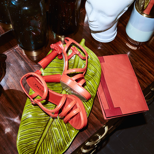 Coral platform stilettos by Chanel are staged on a green leaf tray alongside a matching clutch on a glossy wood surface.