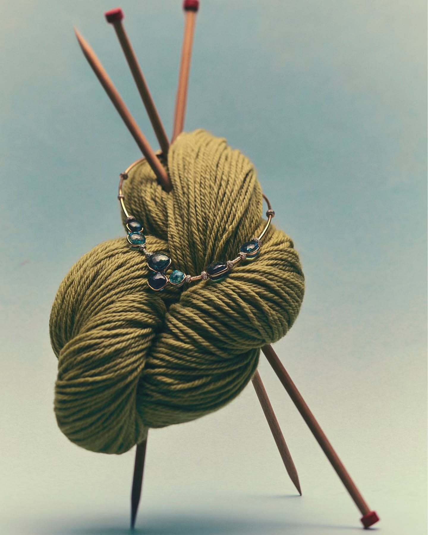 Close-up of olive-green yarn ball pierced by wooden knitting needles, accessorized with a beaded necklace, on a pale blue backdrop.