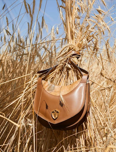 Brown leather hobo bag in a field of wheat stalks, photographed in natural light with a blue sky in the background.