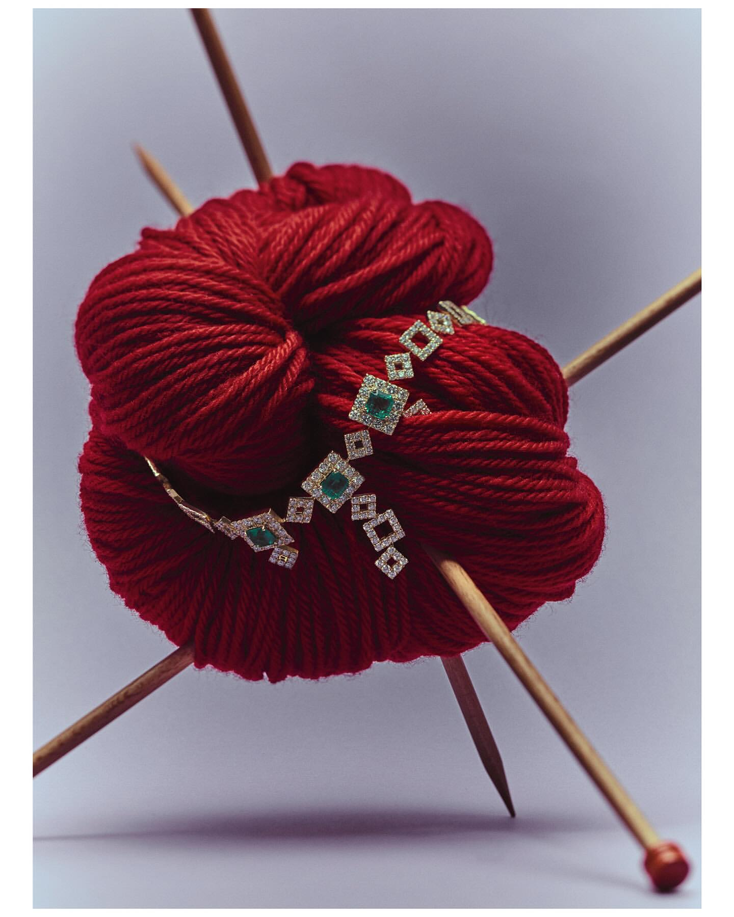 Still life shot of a ball of red yarn with knitting needles and a diamond and emerald necklace on a light background.