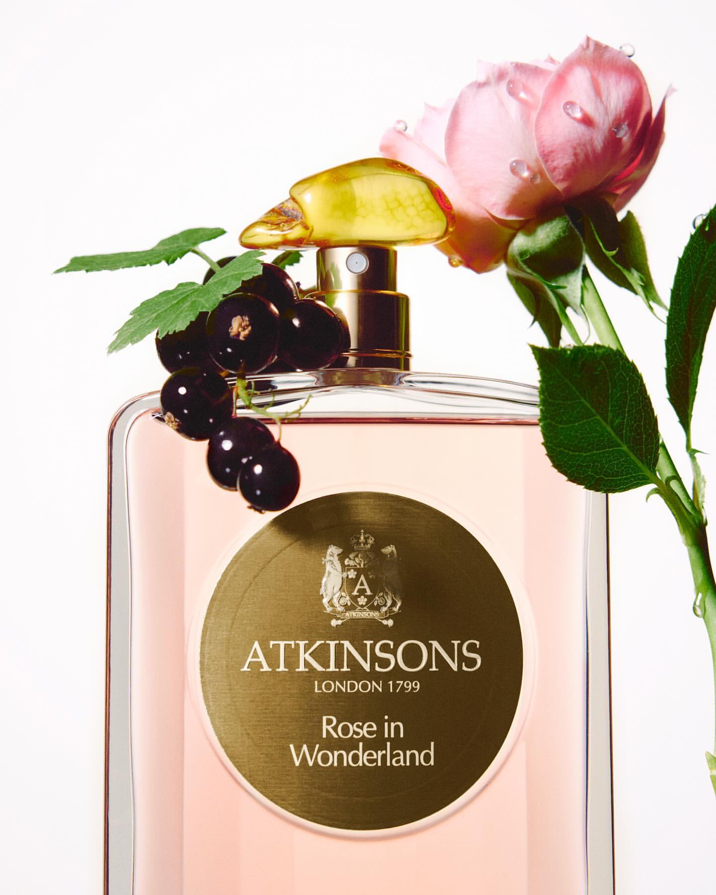 A bottle of Atkinson's Rose in Wonderland perfume is adorned with blackcurrants and a pink rose, set against a clean white backdrop.