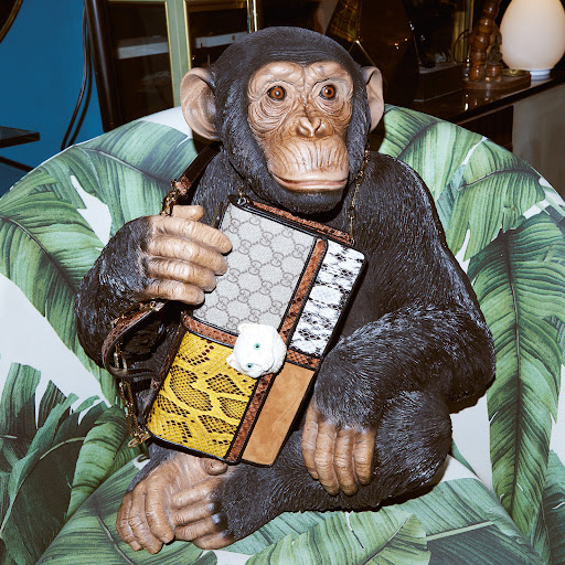 Chimpanzee statue with a purse on a chair with leaf pattern, in an indoor environment with other items.