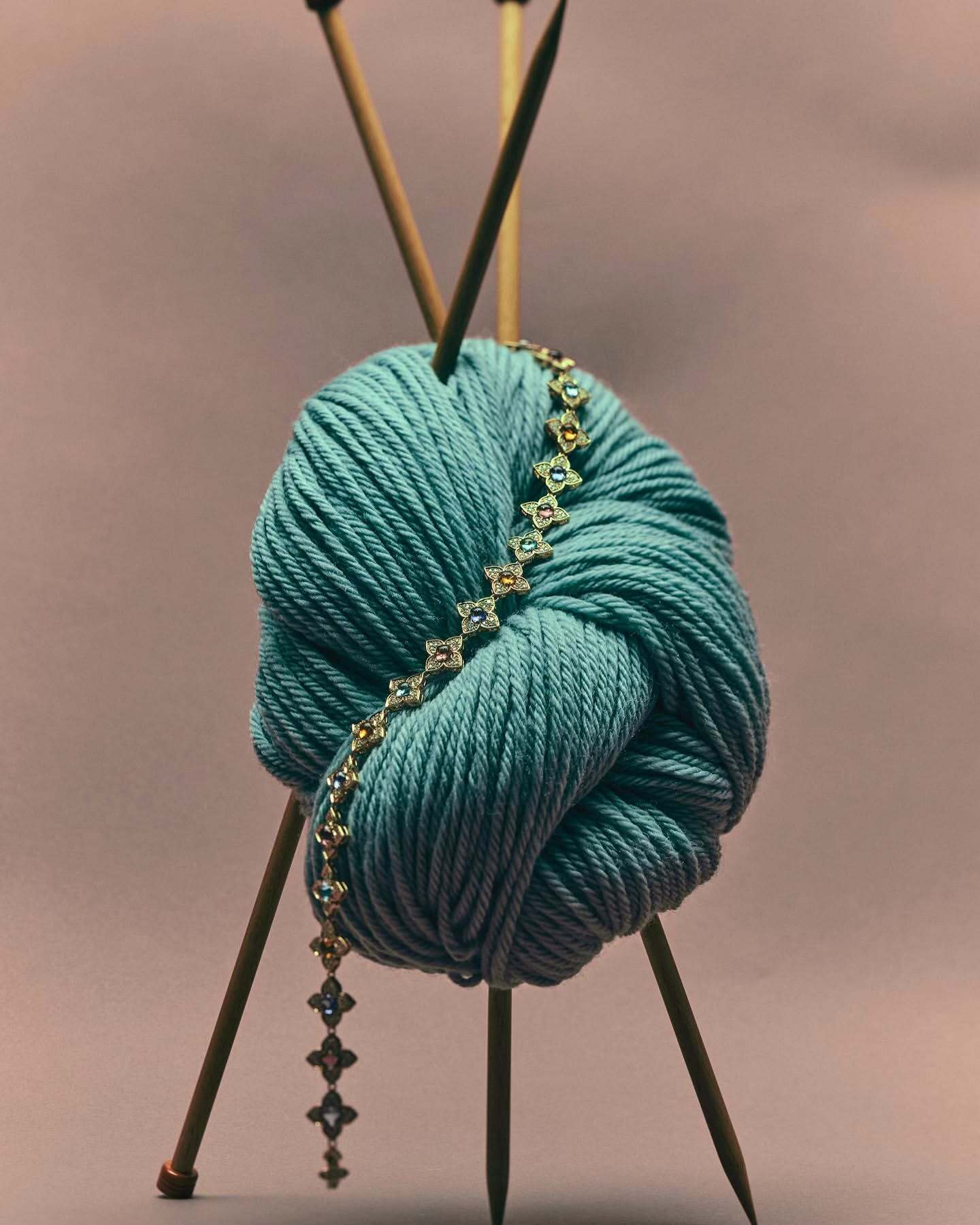 Close-up of a turquoise yarn ball pierced by knitting needles and adorned with a delicate gold bracelet on a mauve backdrop.
