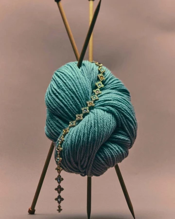 Close-up of a turquoise yarn ball pierced by knitting needles and adorned with a delicate gold bracelet on a mauve backdrop.