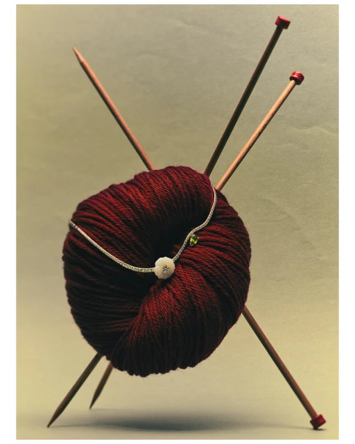 Close-up still life photo of a ball of maroon yarn with decorative string and crossed wooden knitting needles on a neutral background.