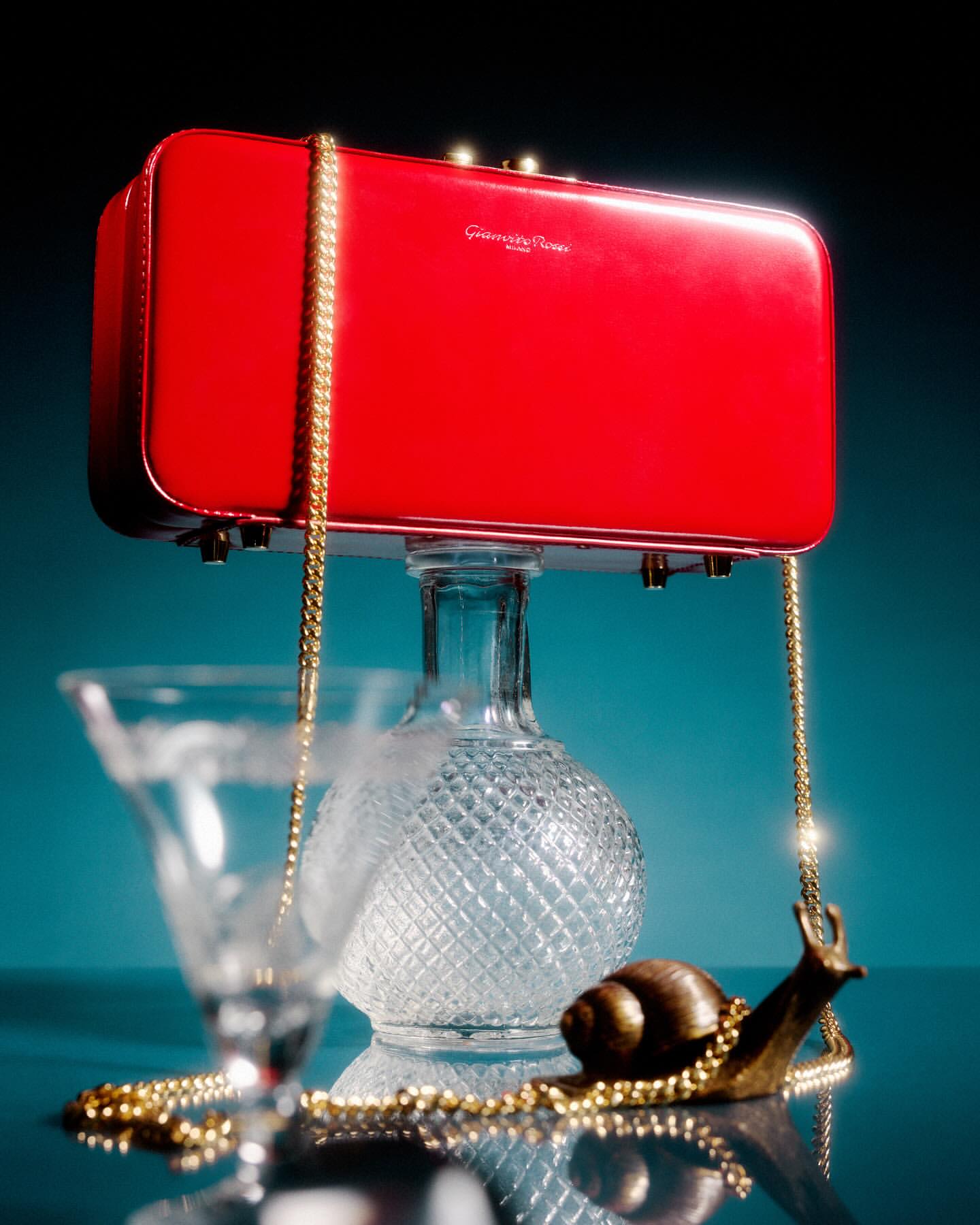 A red handbag with a gold chain strap is showcased on a glass carafe, accompanied by a glass and decorative gold snail.