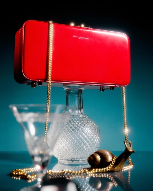 A red handbag with a gold chain strap is showcased on a glass carafe, accompanied by a glass and decorative gold snail.